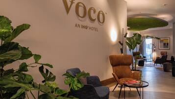 Un moderno vestíbulo de hotel con un gran letrero que dice 'Voco An IHG Hotel'. Hay varias zonas de estar con sillas y mesas, y grandes plantas verdes distribuidas por el espacio.