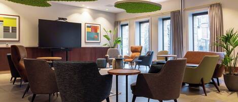 A modern hotel lobby with a flat-screen TV, comfortable seating, large windows, and a ceiling with green accents.
