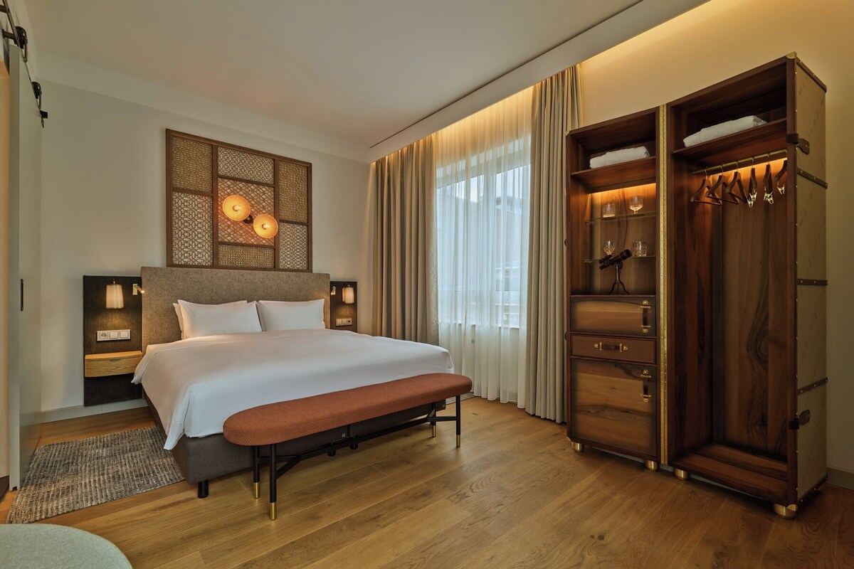A modern hotel room with a large bed, a bench, a wooden wardrobe, and a decorative wall panel.