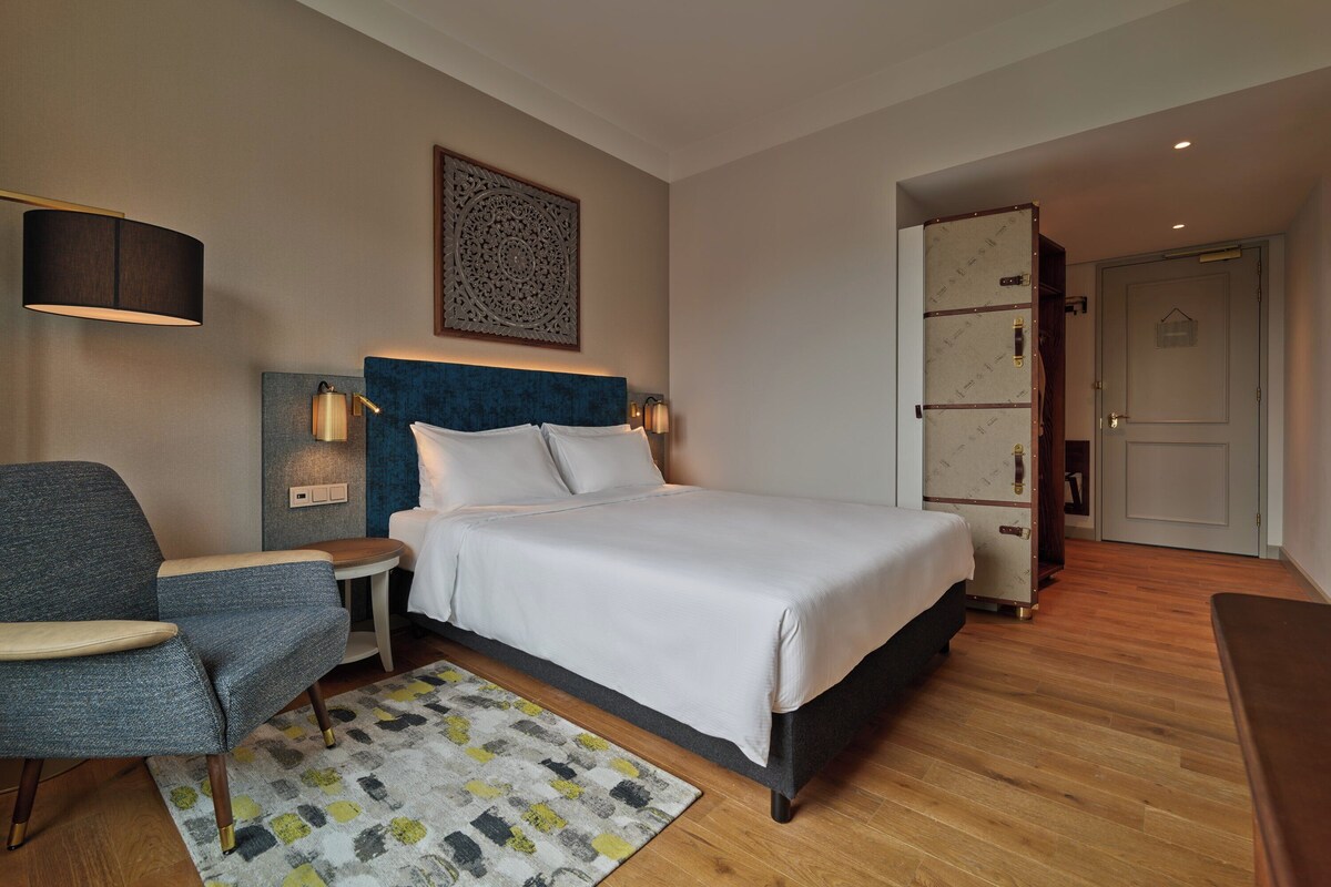 A modern hotel room with a large bed, a blue upholstered chair, a bedside table with a lamp, and a patterned rug.