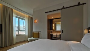 A hotel room with a large bed, a TV, a window with curtains, a bathroom with a mirror, and a light fixture. - voco The Hague by IHG (The Hague)