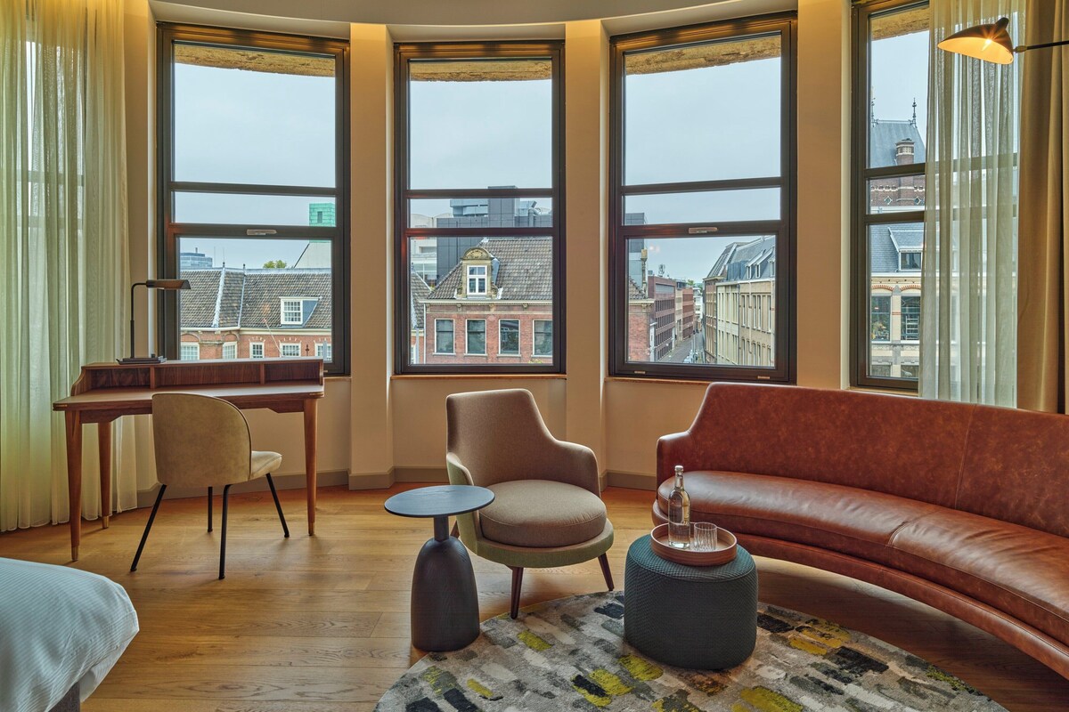 A modern living room with a brown leather sofa, a round coffee table, and a desk with a chair. Large windows offer a view of buildings outside.