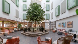 A spacious indoor area with a central tree, seating arrangements, and artwork on the walls. - voco The Hague by IHG (The Hague)