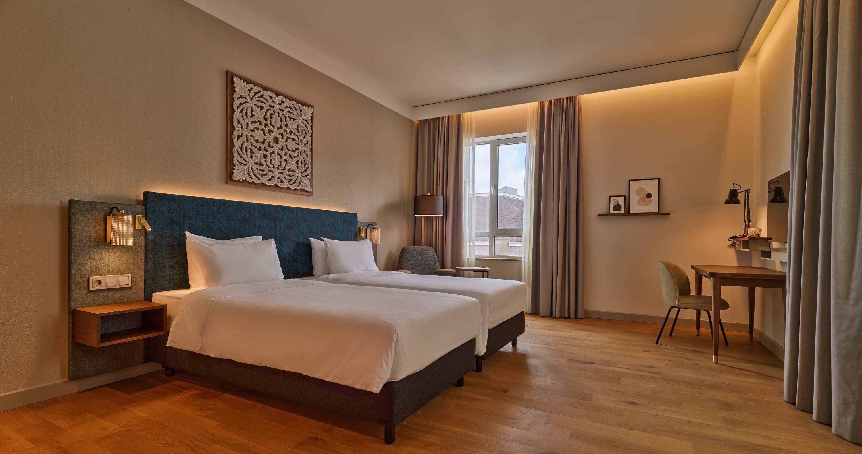 A hotel room with two beds, a desk, a chair, a lamp, and a window with curtains.