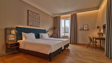 A hotel room with two beds, a desk, a chair, a lamp, and a window with curtains.