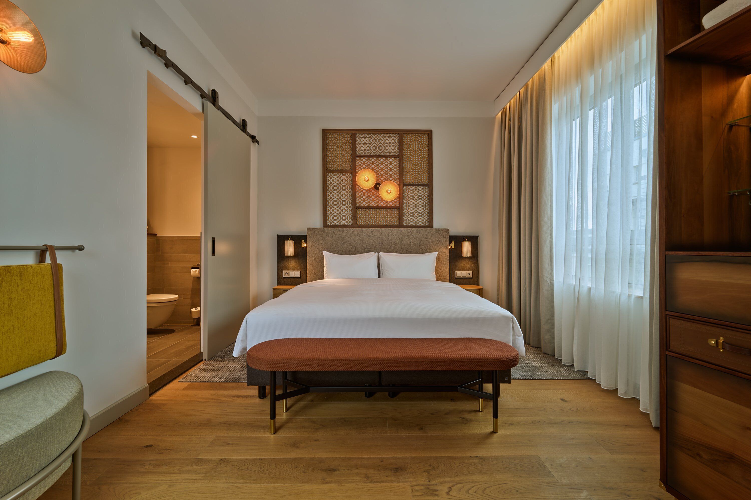 Habitación de hotel con una cama grande, cabecero de madera, lámparas de noche, un banco y vistas de la ciudad a través de cortinas translúcidas.