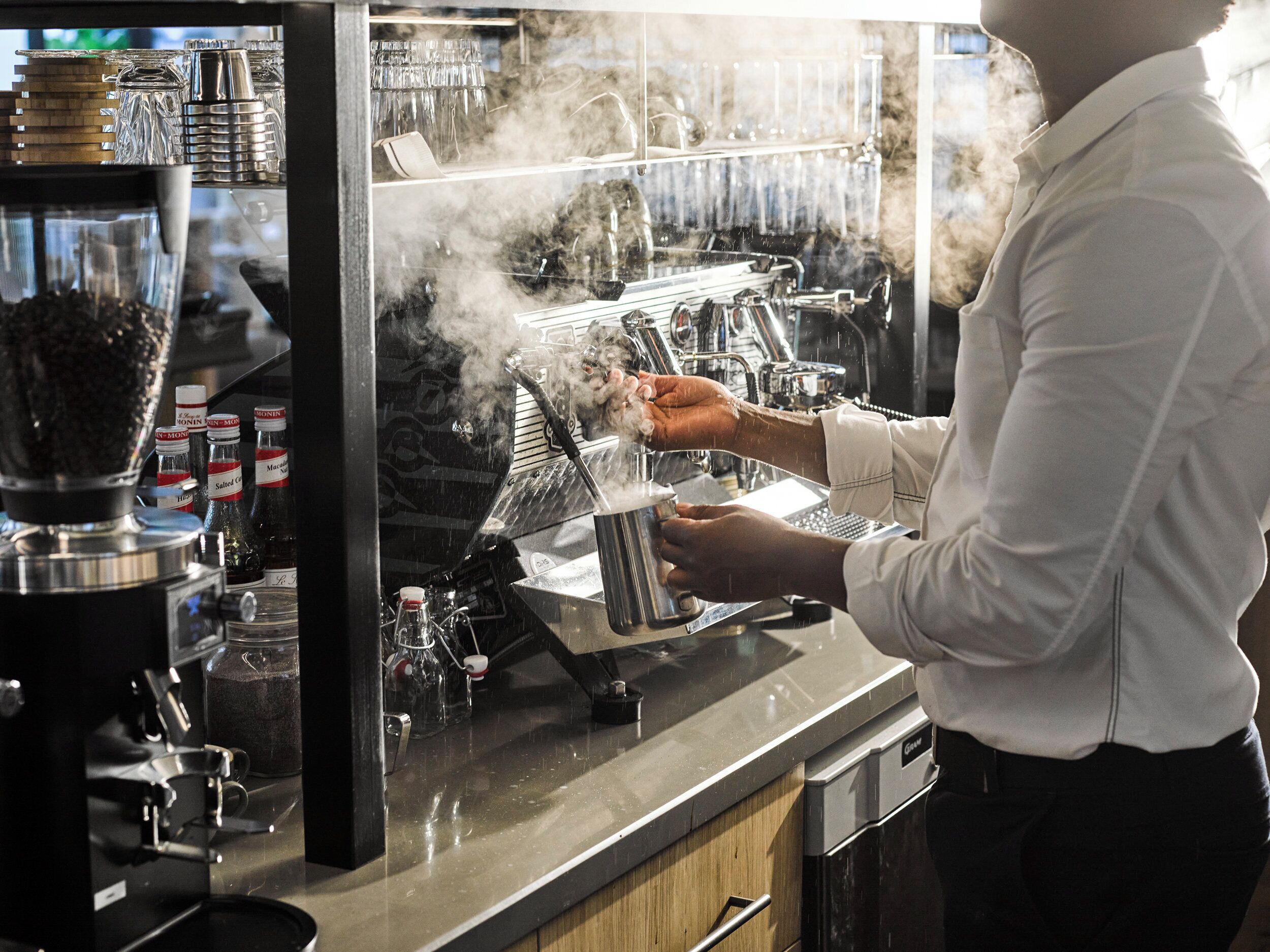 Ein Barista bereitet ein Getränk mit einer Dampfdüse, einer Kaffeemaschine sowie verschiedenen Kaffeekapseln und Behältern zu.