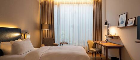Une chambre d’hôtel avec un lit, un bureau, une chaise, une lampe et une fenêtre avec des rideaux.