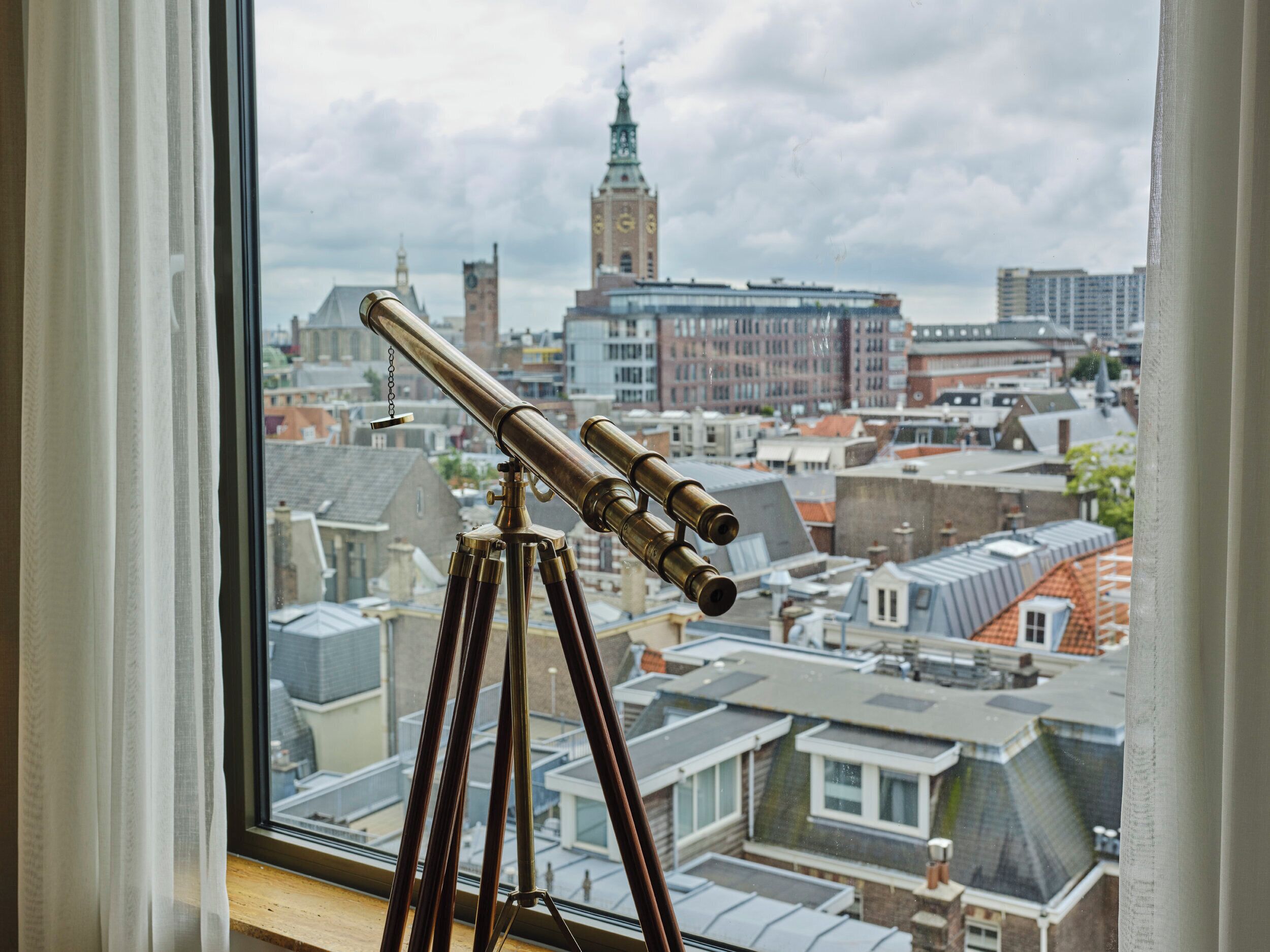 A telescope on a tripod by a window overlooking a cityscape with historic buildings and a clock tower.