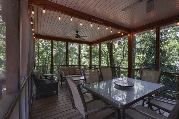 A covered outdoor seating area with a table and chairs, surrounded by large windows offering a view of the forest.