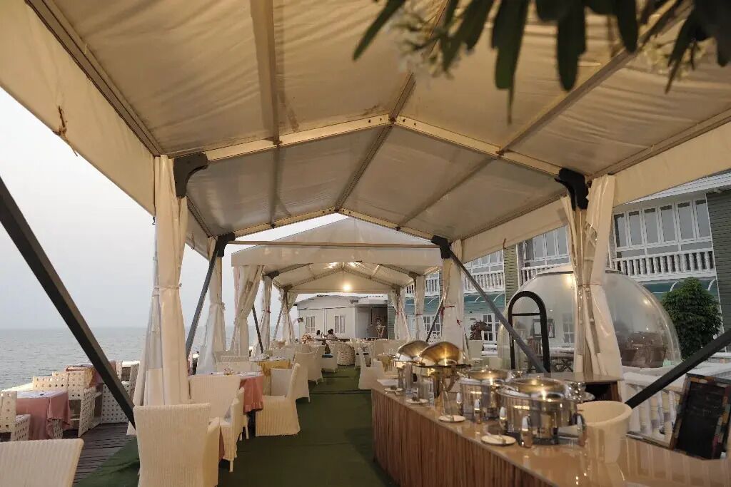 Un espace de restauration aménagé sous une tente, avec des tables et des chaises, un bar équipé de verres et d’ustensiles, et une vue sur la mer.