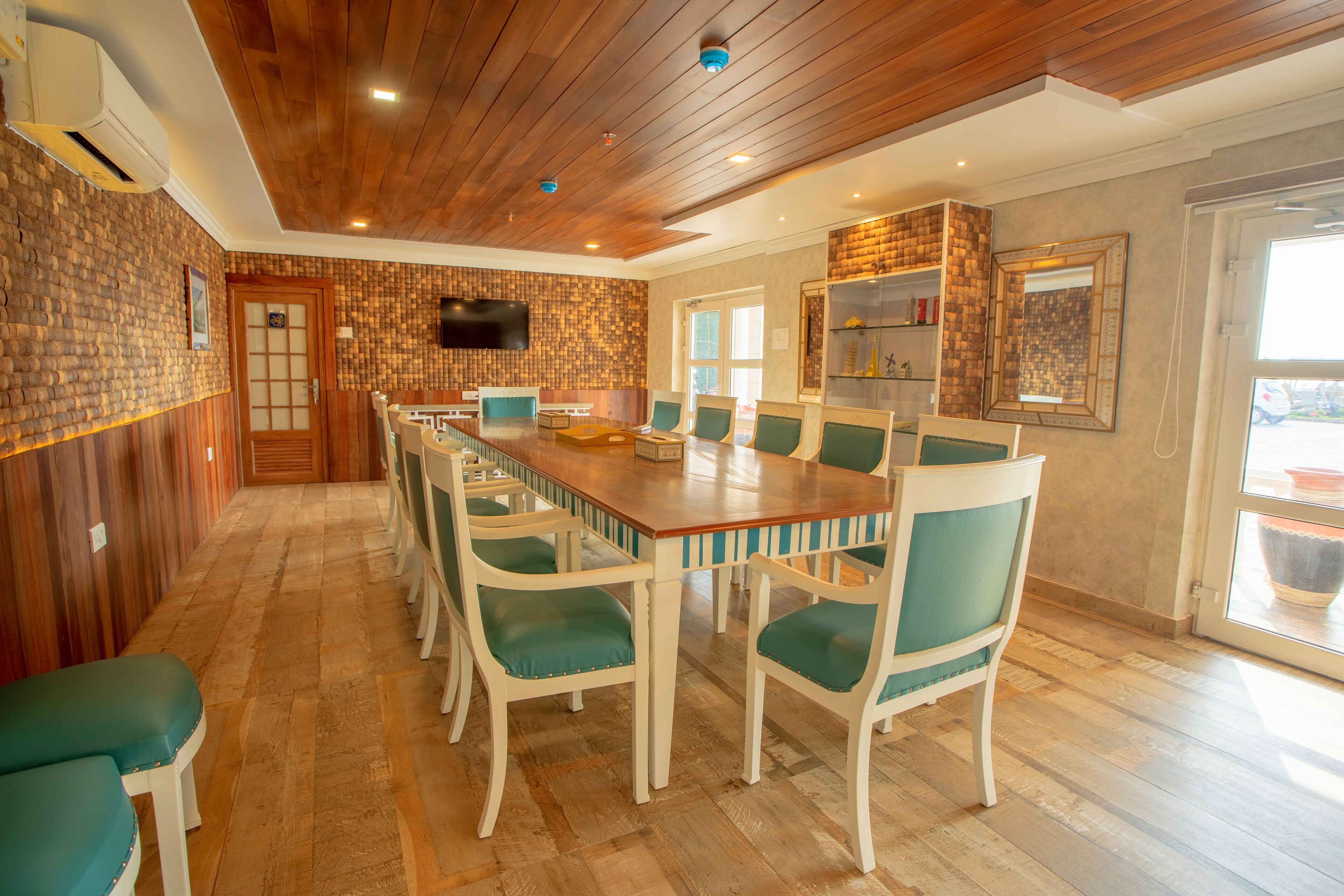 A dining area with a long wooden table and teal chairs, a TV mounted on the wall, and a brick-patterned wall.