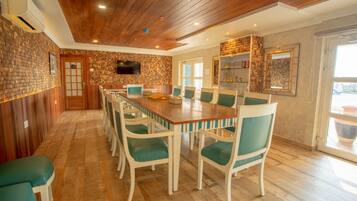 Un coin repas avec une longue table en bois et des chaises de couleur turquoise, un téléviseur fixé au mur et un mur décoré d’un motif en briques.
