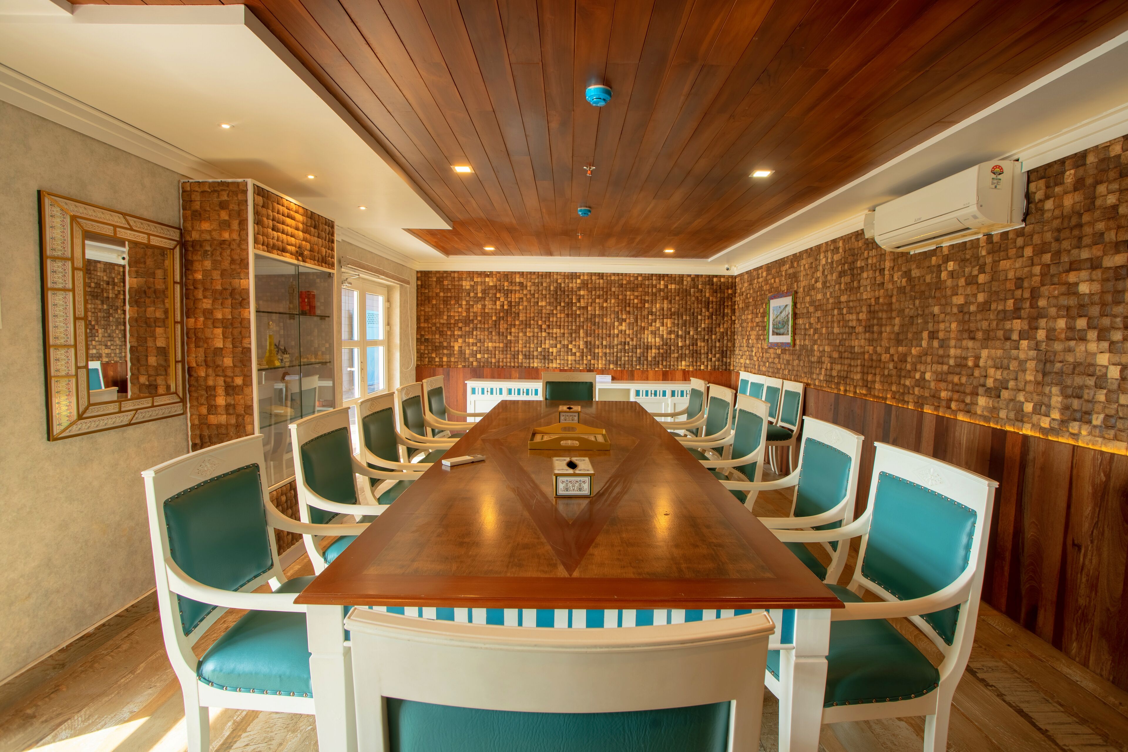 Ein Konferenzraum mit einem langen Holztisch, weißen Stühlen mit türkisfarbenen Kissen und einer Wand mit Backsteinmuster.