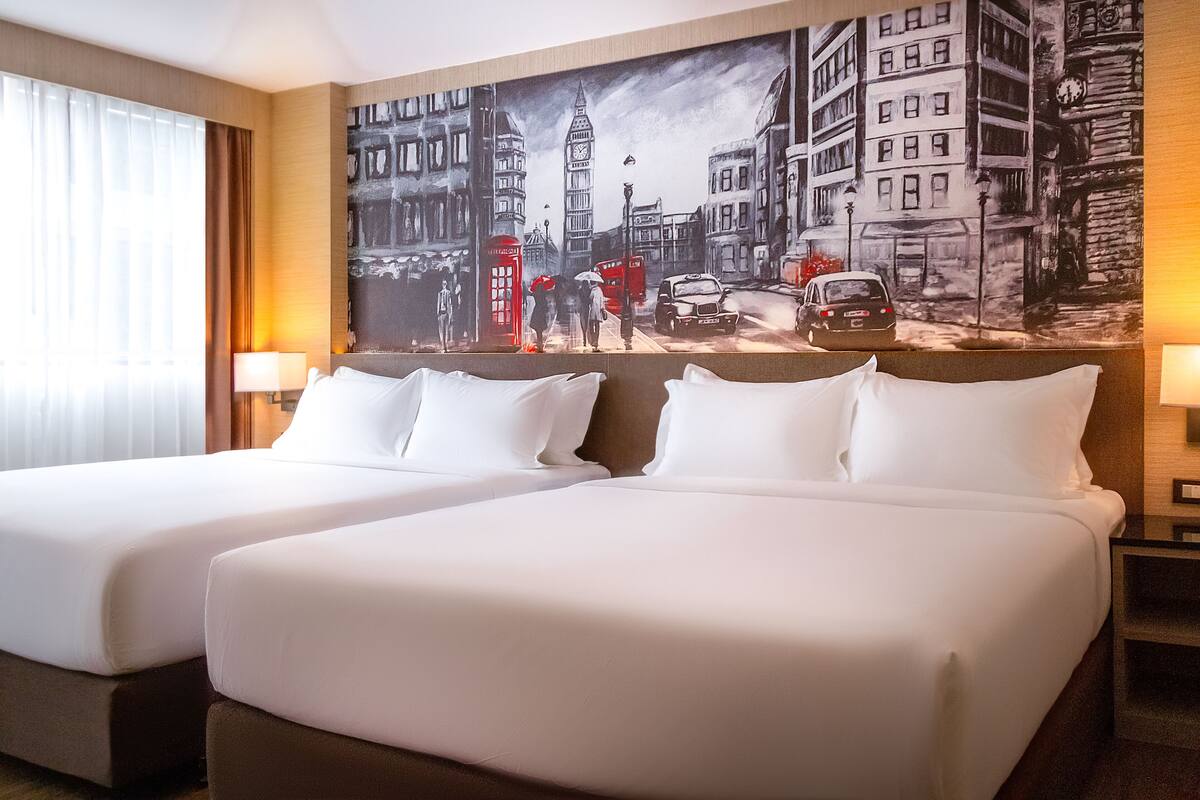 A hotel room with two beds, a large cityscape mural on the wall, and a window with curtains.