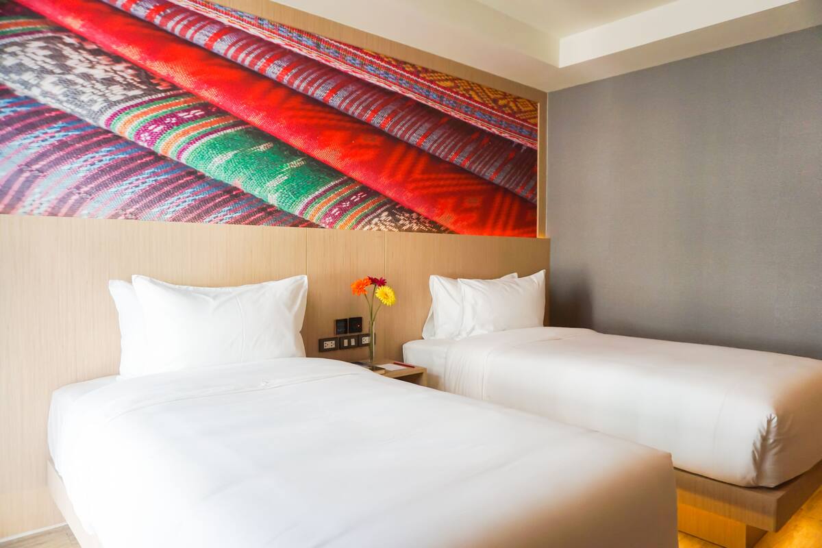 A hotel room with two beds, a wall-mounted tapestry, and a small table with a vase of flowers.