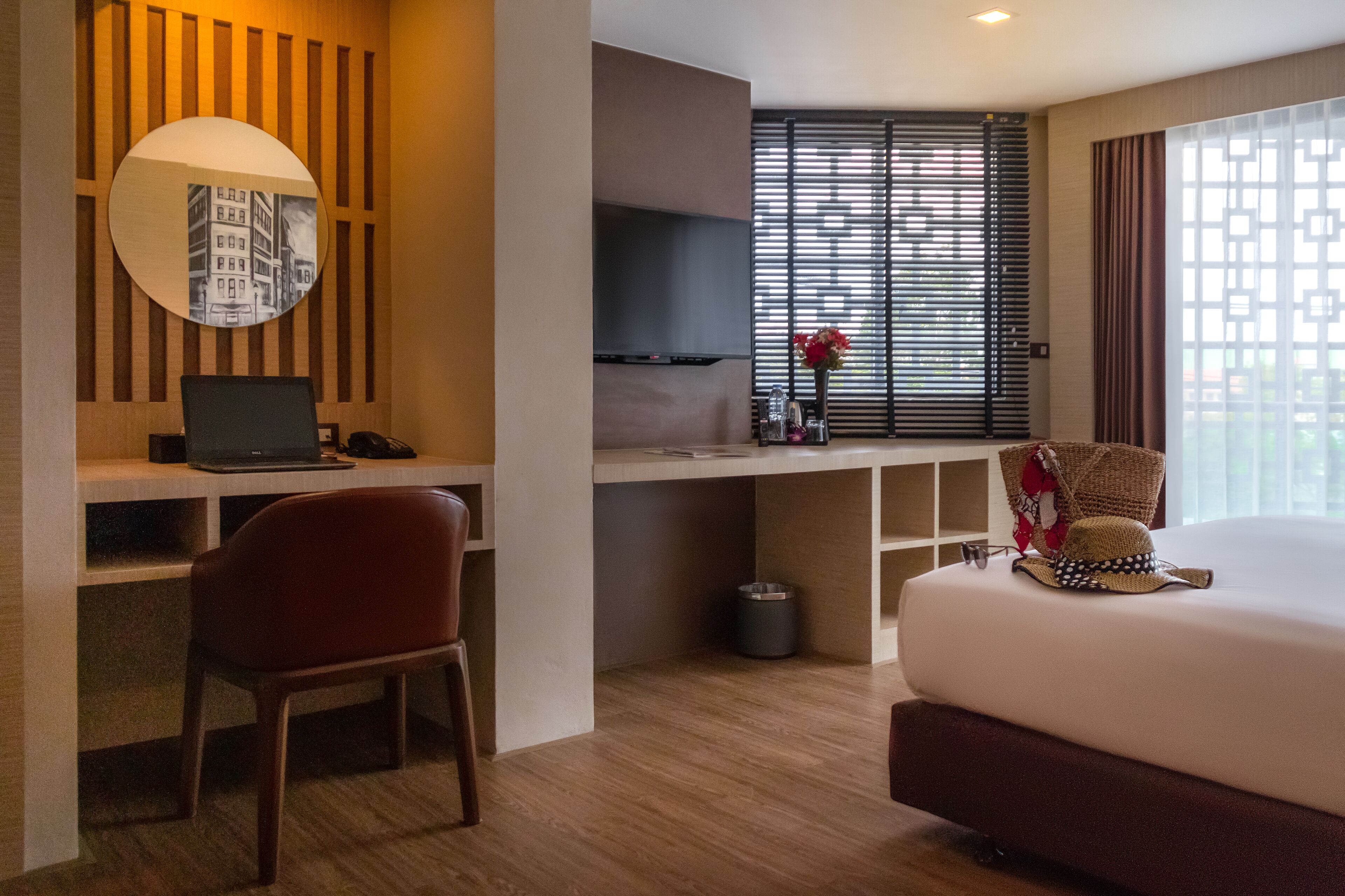 A hotel room with a bed, a desk with a laptop, a chair, a TV, and a window with blinds.