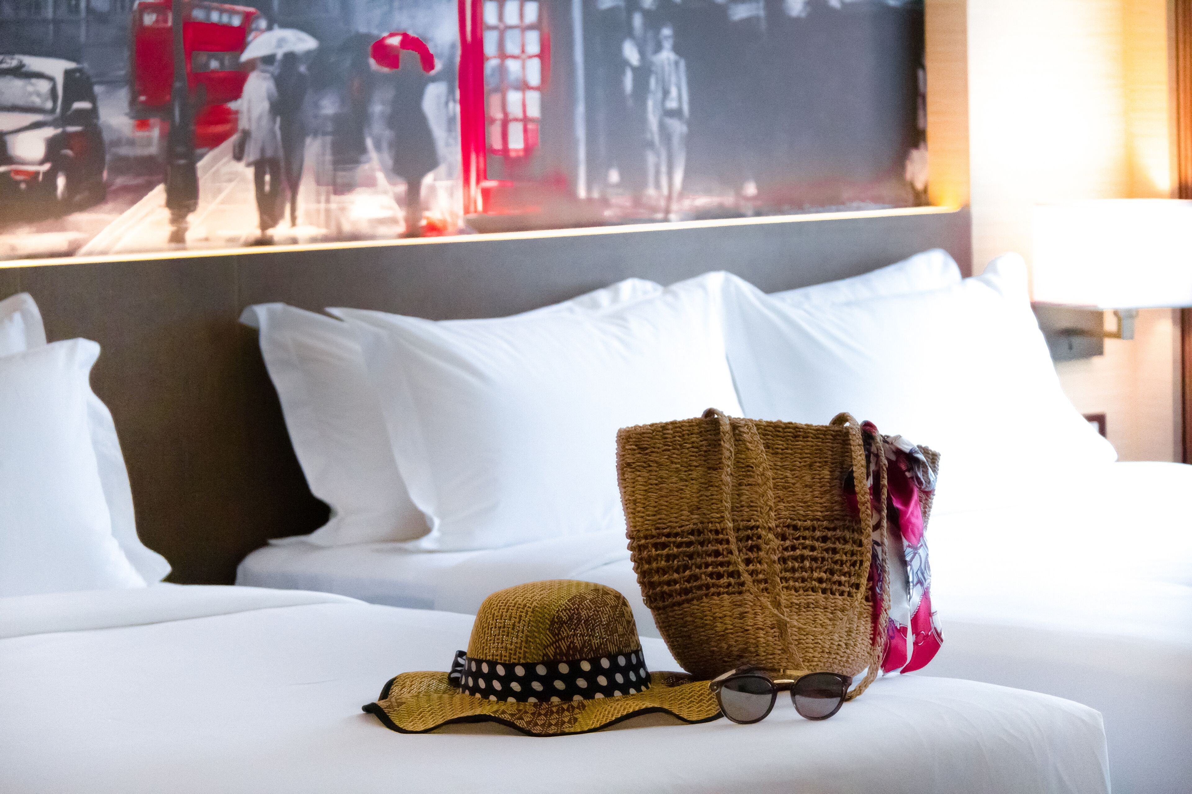 Una cama con ropa de cama blanca, un bolso tejido, un sombrero de paja y unas gafas de sol.