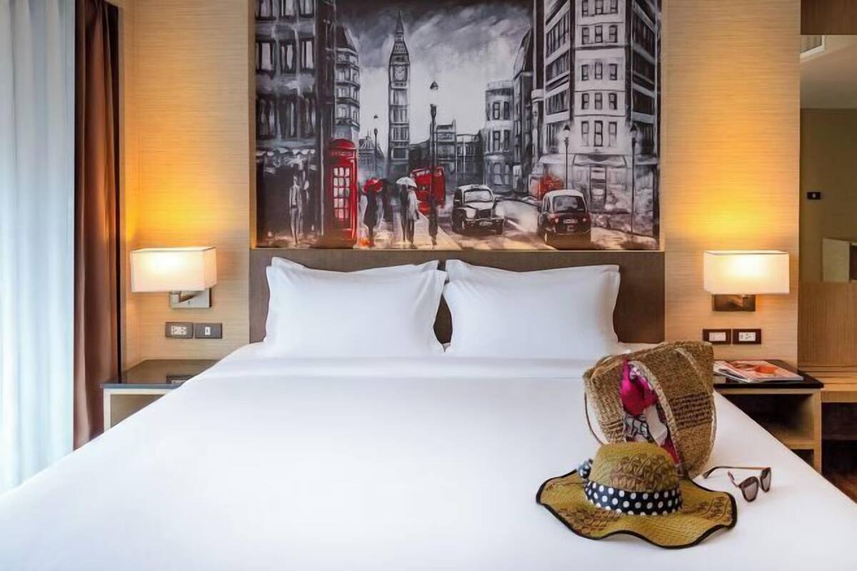 A hotel room with a large bed, two bedside lamps, a painting of a cityscape, and a decorative hat on the bed.