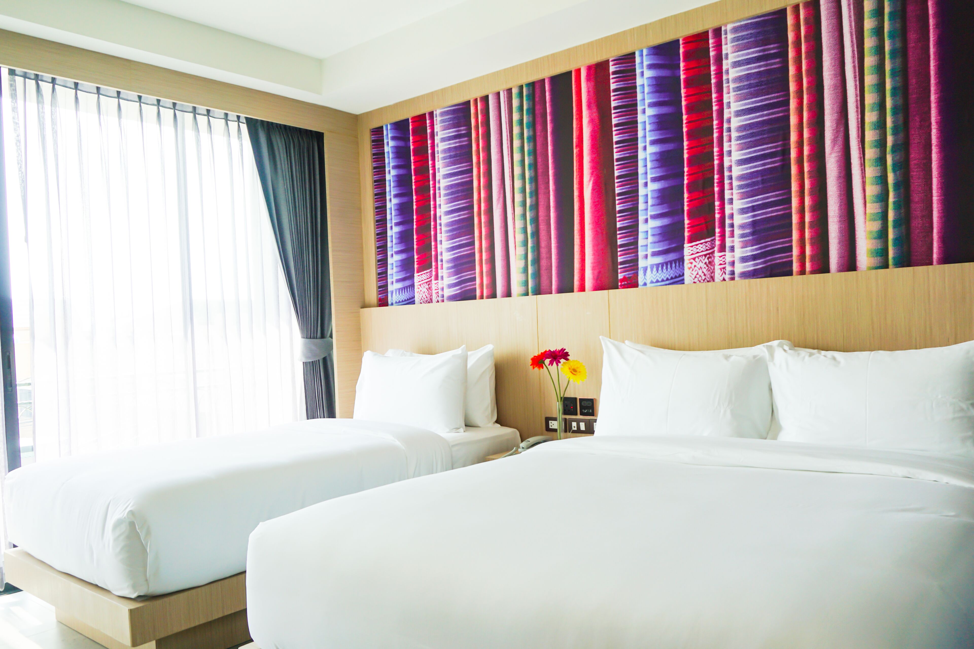 A hotel room with two beds, a large window with curtains, and a colorful tapestry on the wall.