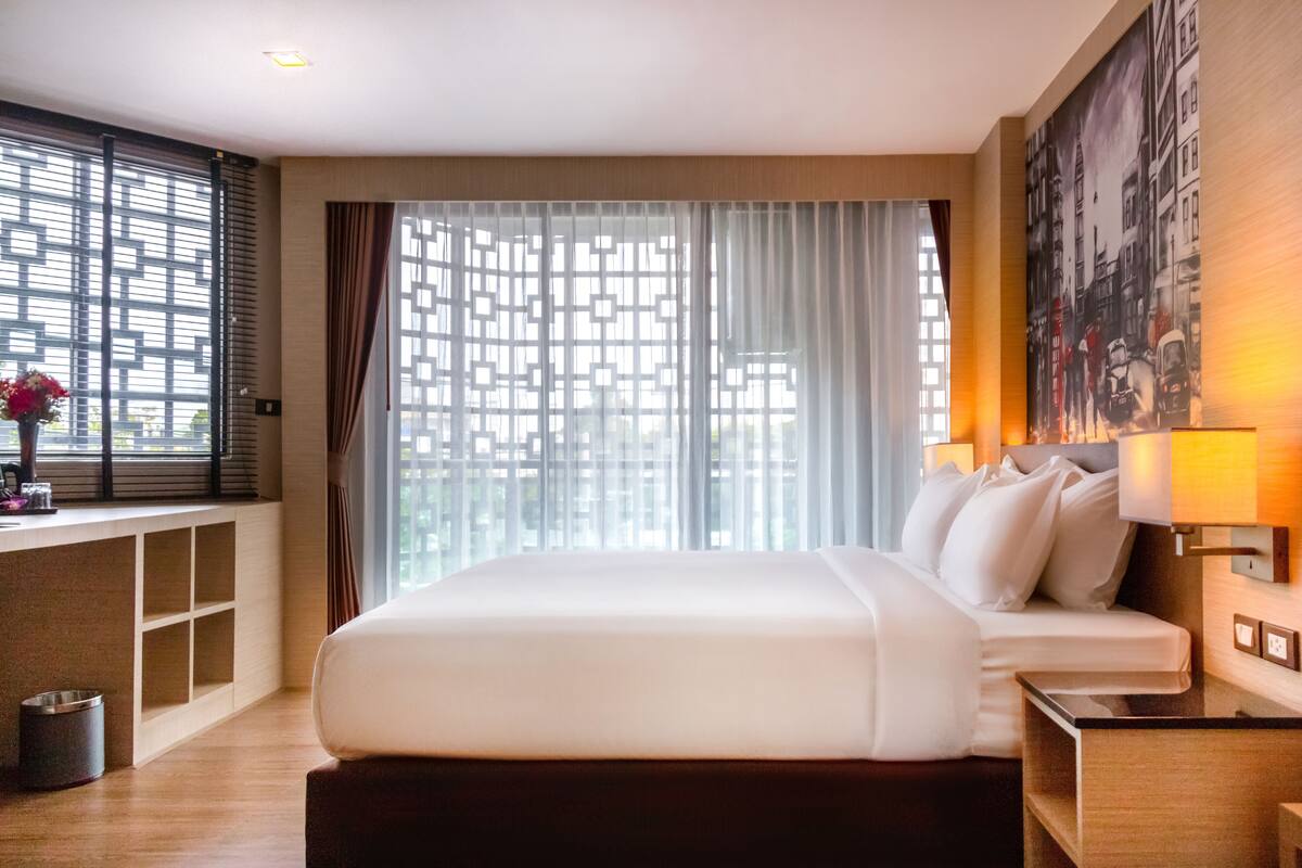 A hotel room with a large bed, a bedside table with a lamp, a window with blinds, a view of the city, and a painting on the wall.