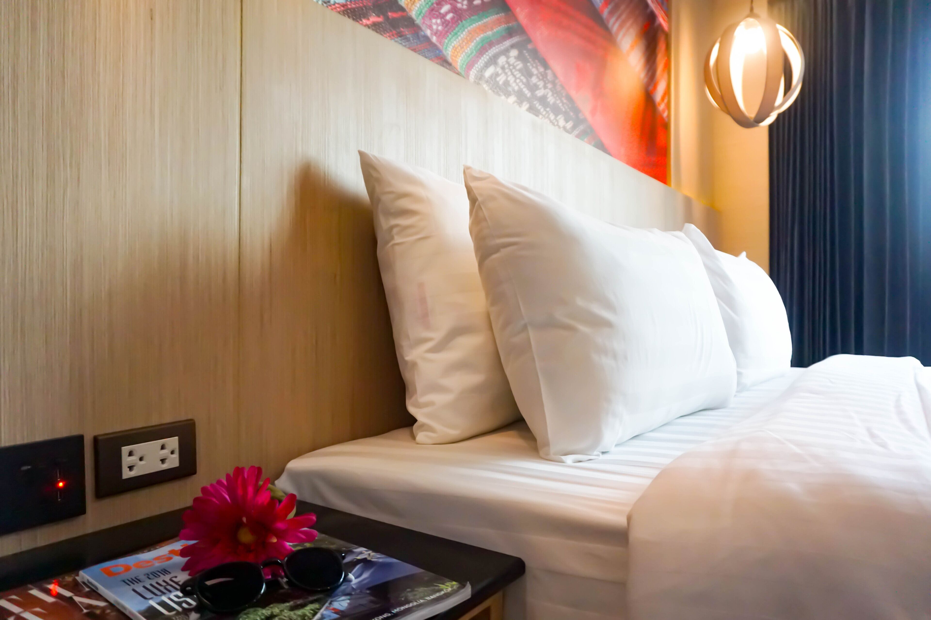 A hotel room with a wooden headboard, white bedding, a magazine, sunglasses, and a pink flower on the nightstand.