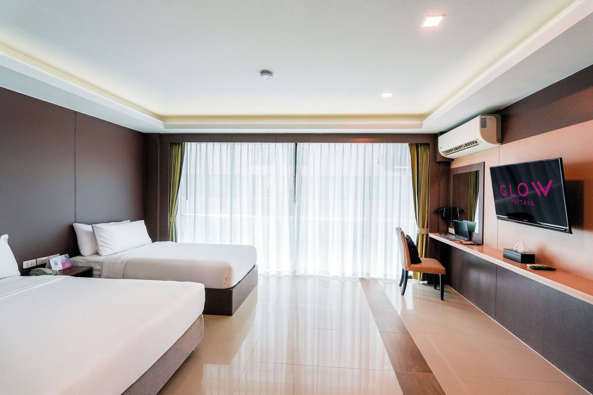 A hotel room with two beds, a TV, a desk, and a large window with curtains.