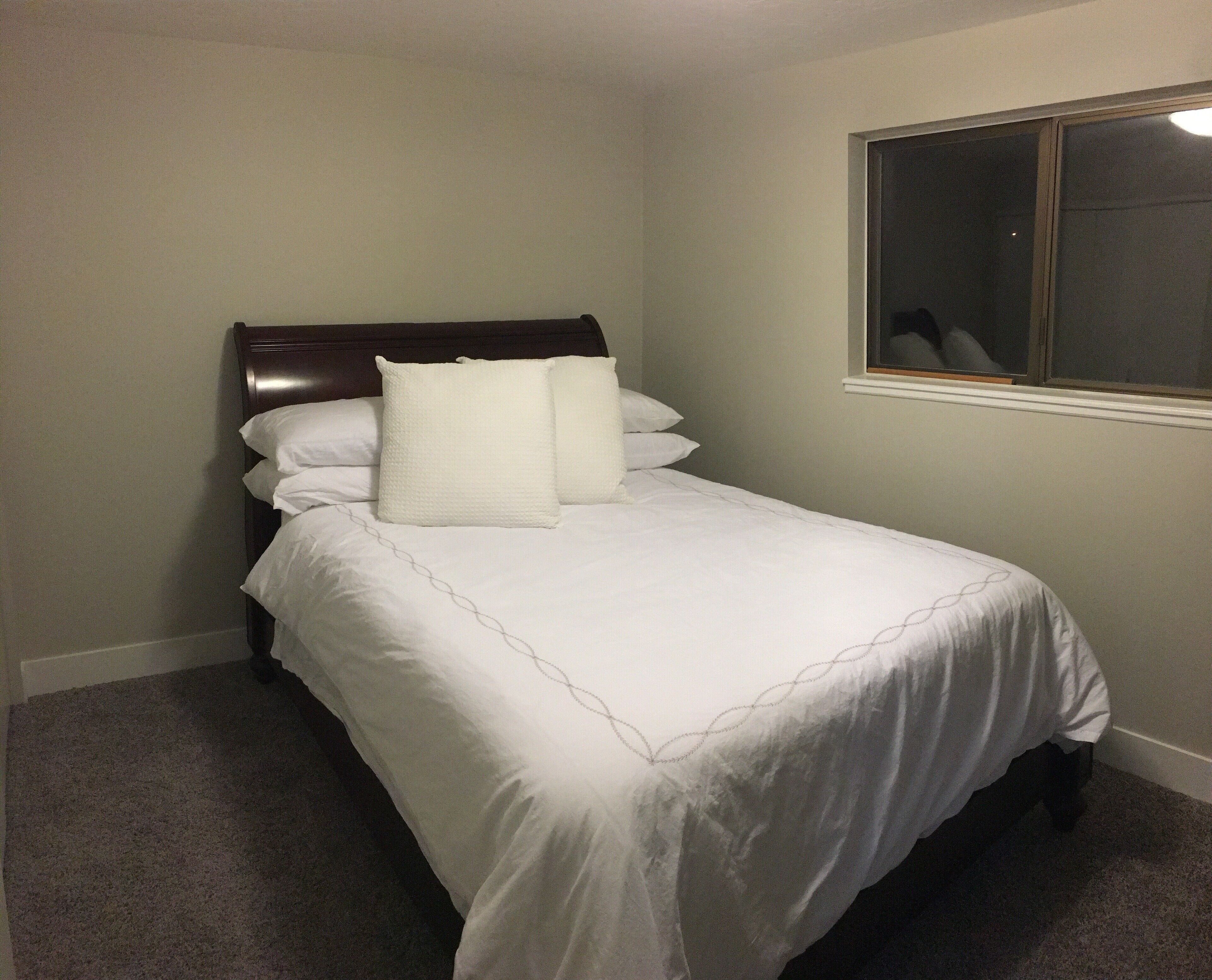 A neatly made bed with white bedding and pillows, a dark headboard, and a window with a view of the outside.