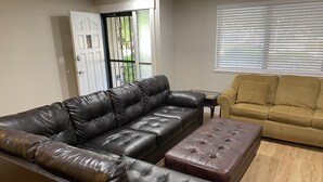 Ein Wohnzimmer mit einem dunkelbraunen Ledersofa, einem kleineren braunen Hocker und einem gelben Sofa. Im Hintergrund ist ein Fenster mit Jalousien zu sehen.