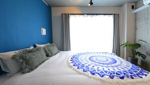 A neatly made bed with a blue and white patterned blanket, a potted plant, and a window with curtains. - Luana Shibuya (Tokyo)