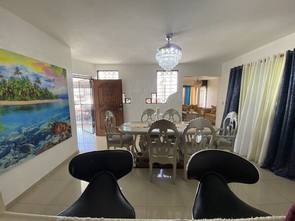 A dining area with a chandelier, a painting of a tropical scene, and a view of the living room.