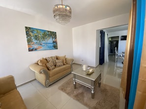 A living room with a sofa, a coffee table, a painting of a tropical beach, and a chandelier.