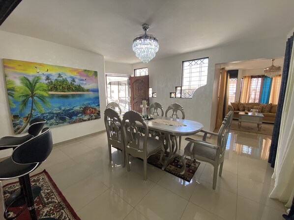 Un comedor con una mesa y sillas, una araña, un cuadro de una escena tropical y vistas a una sala de estar.