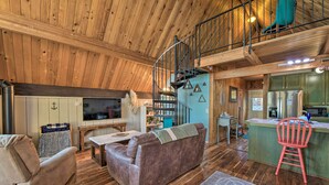 A spacious living area with a wooden ceiling, a sofa, a coffee table, and a TV. There is a kitchen area with a refrigerator and a sink.