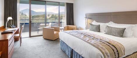 A hotel room with a large bed, a desk, a chair, and a balcony with a view of the mountains.