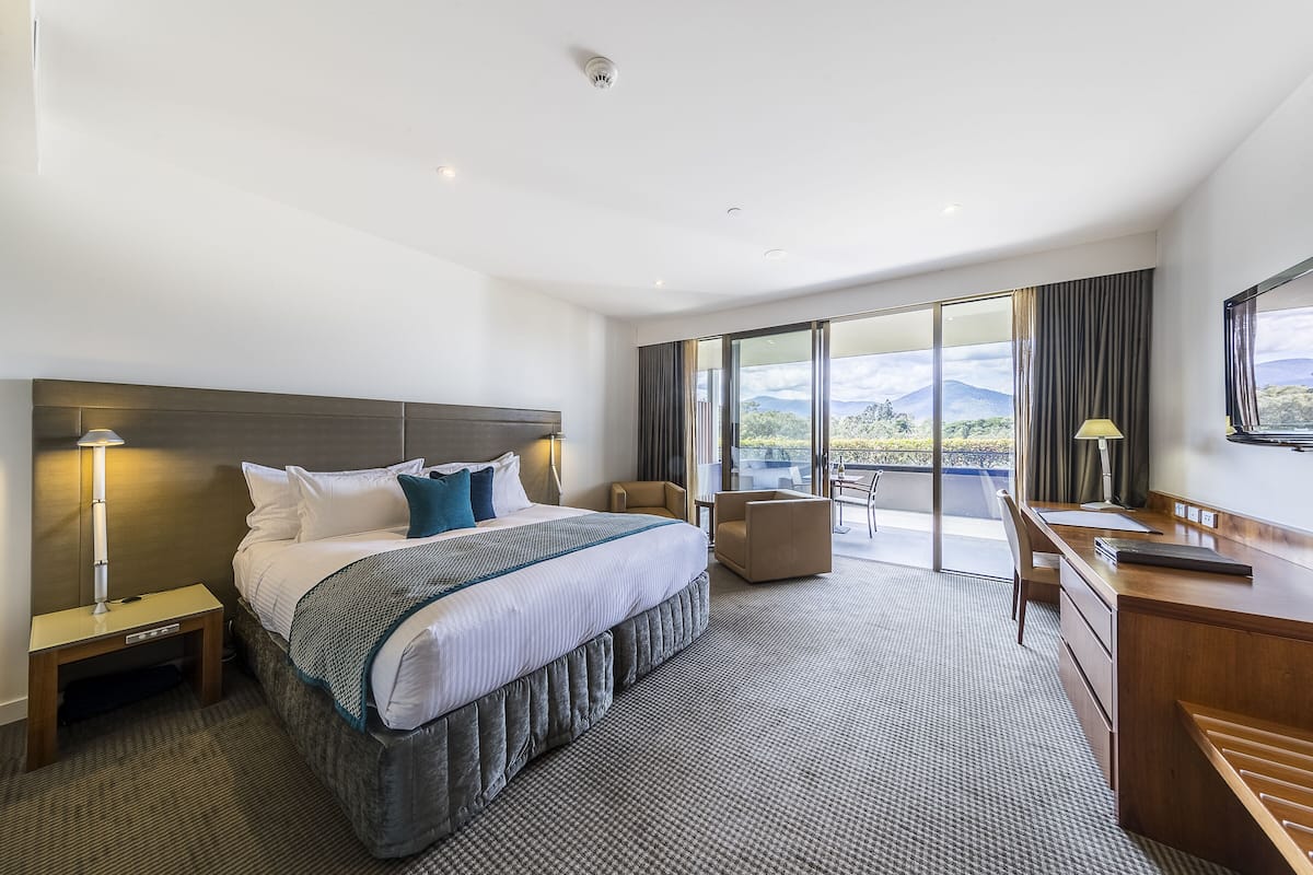 A modern hotel room with a large bed, a desk, and a view of the mountains through the windows.