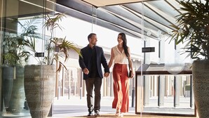 Un couple se tenant la main dans un hall d’entrée moderne, agrémenté de grandes plantes en pot et doté d’une entrée en verre.