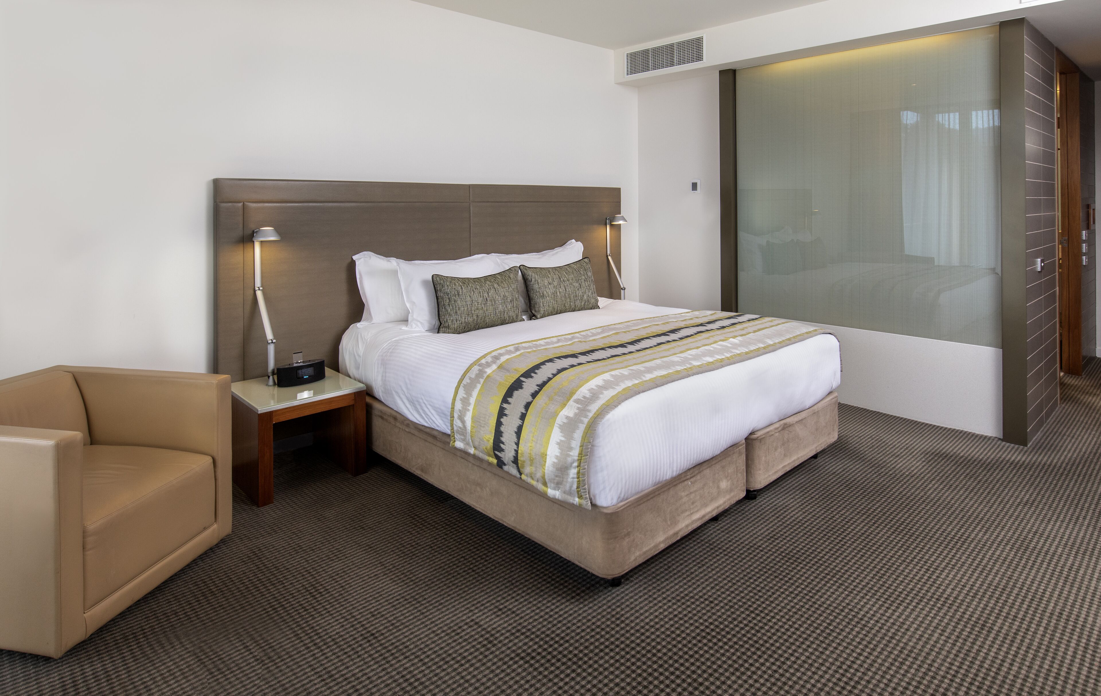 A hotel room with a large bed, a bedside table with a lamp, a chair, and a sliding glass door to the bathroom.