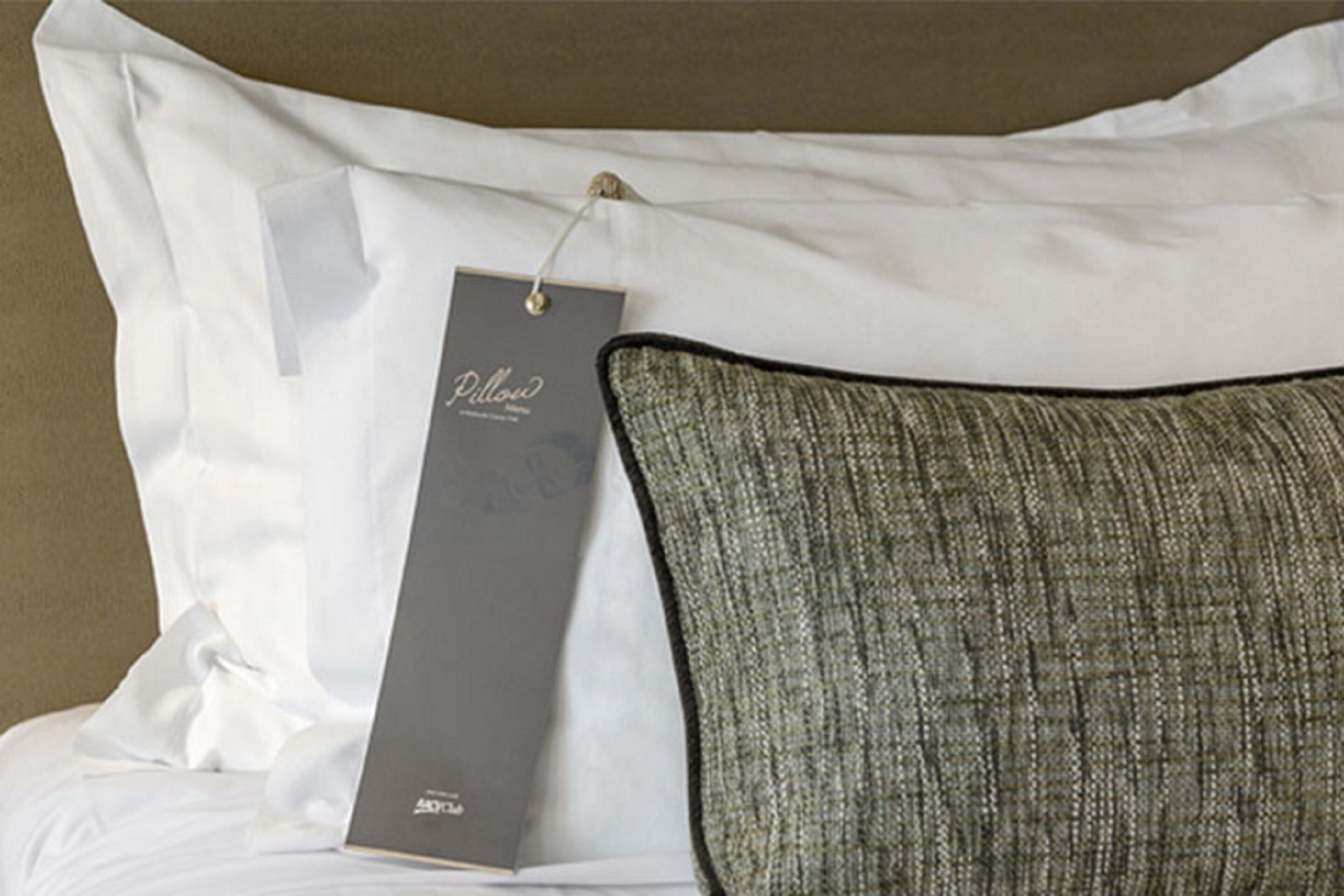 A bed with white pillows and a grey pillow with a tag.