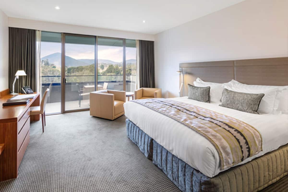 A hotel room with a large bed, a desk, a chair, and a balcony with a view of the mountains.