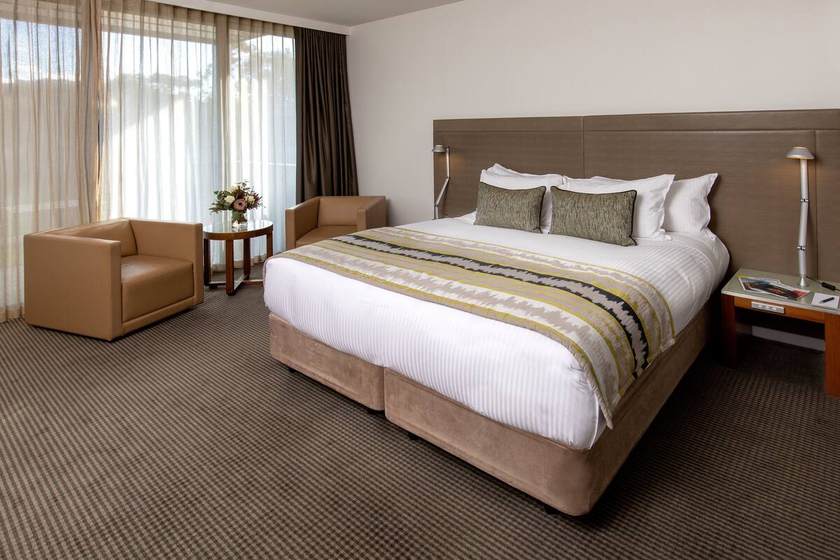 A hotel room with a large bed, two chairs, a small table with a vase, and a nightstand with a lamp.