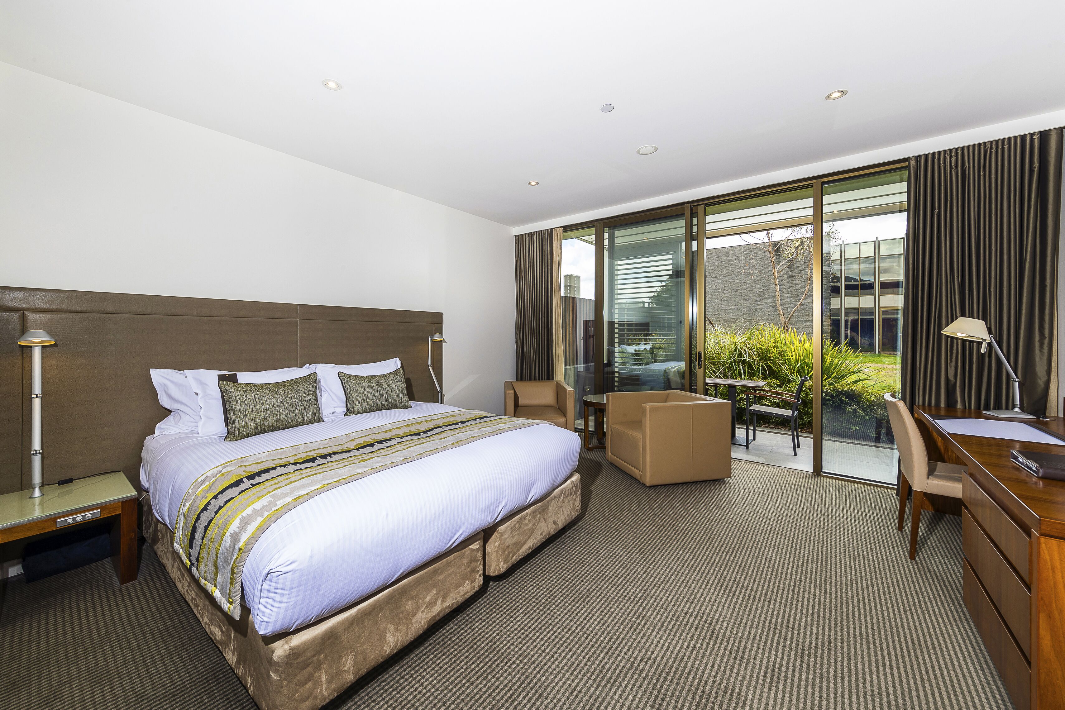 A modern hotel room with a large bed, a desk, a chair, and a balcony with a view of the city.