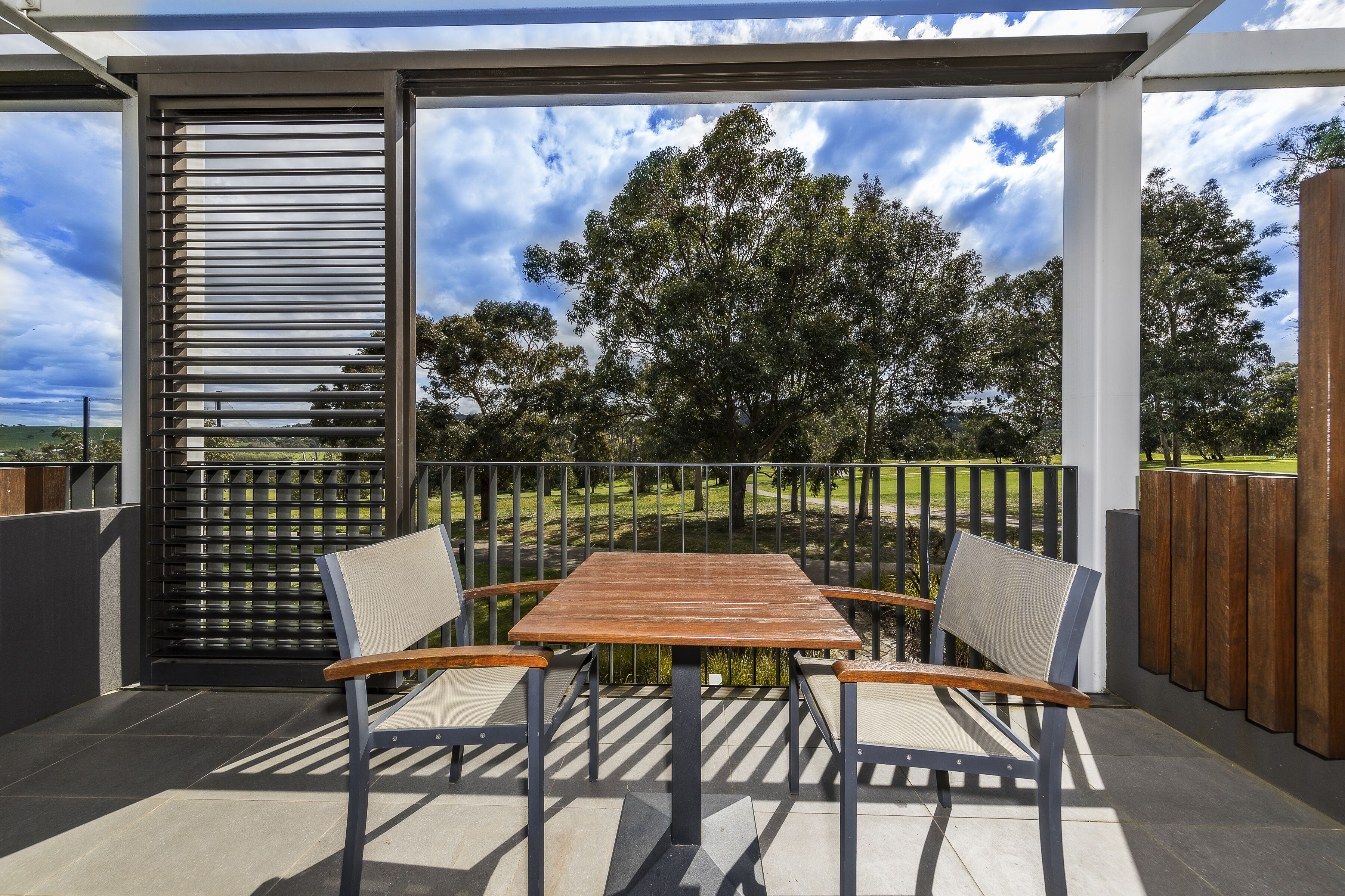 An outdoor patio with a wooden table and chairs, a metal railing, and a view of trees and a cloudy sky.