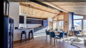 A modern kitchen with wooden beams, a dining area with blue chairs, and large windows offering a mountain view. - Prestigious ski-in/ski-out apartment with panoramic view (Huez)