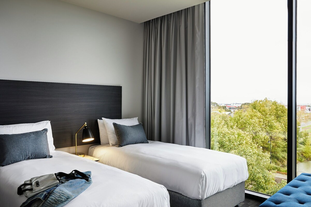 Habitación de hotel con dos camas, un ventanal con vistas a zonas verdes y edificios, un cabecero oscuro y un sillón tapizado en azul.