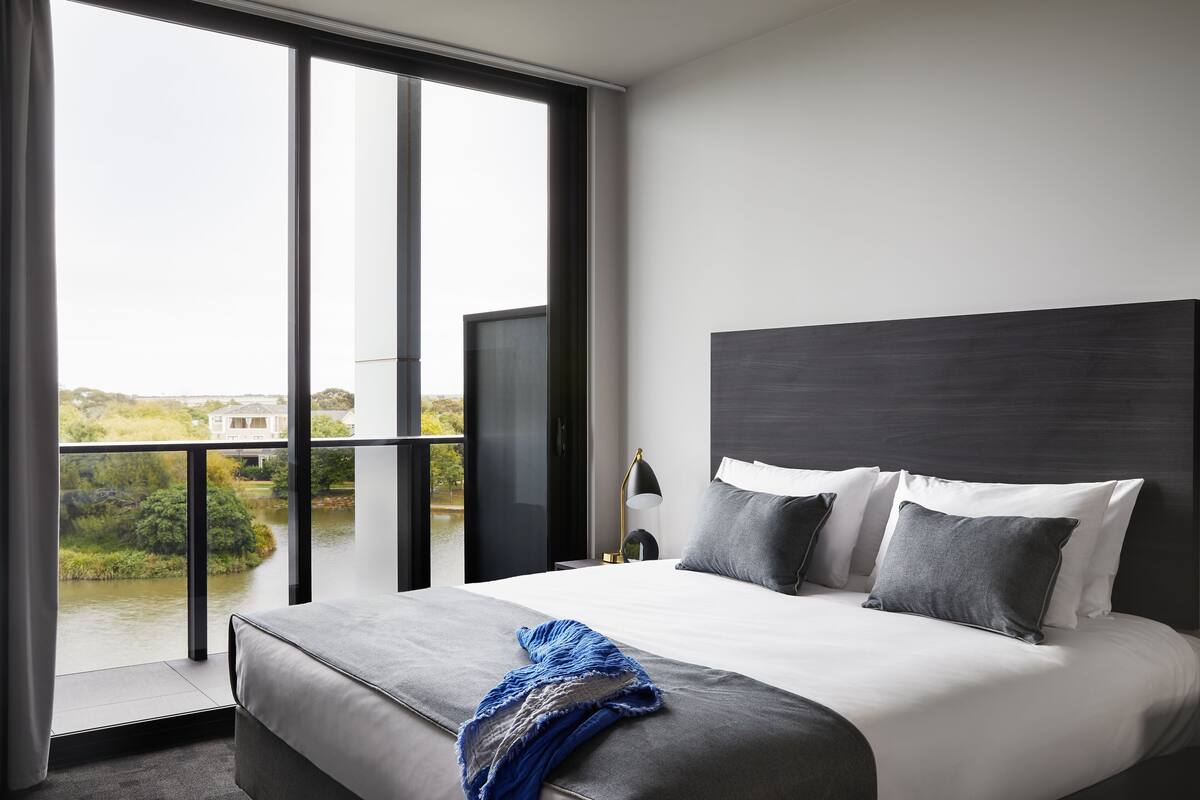 A modern hotel room with a large bed, a nightstand, a lamp, and a view of a river and buildings through large windows.