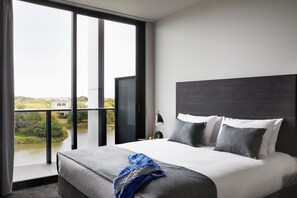 A modern hotel room with a large bed, a nightstand, a lamp, and a view of a river and buildings through large windows. - Punthill Caroline Springs (Caroline Springs)