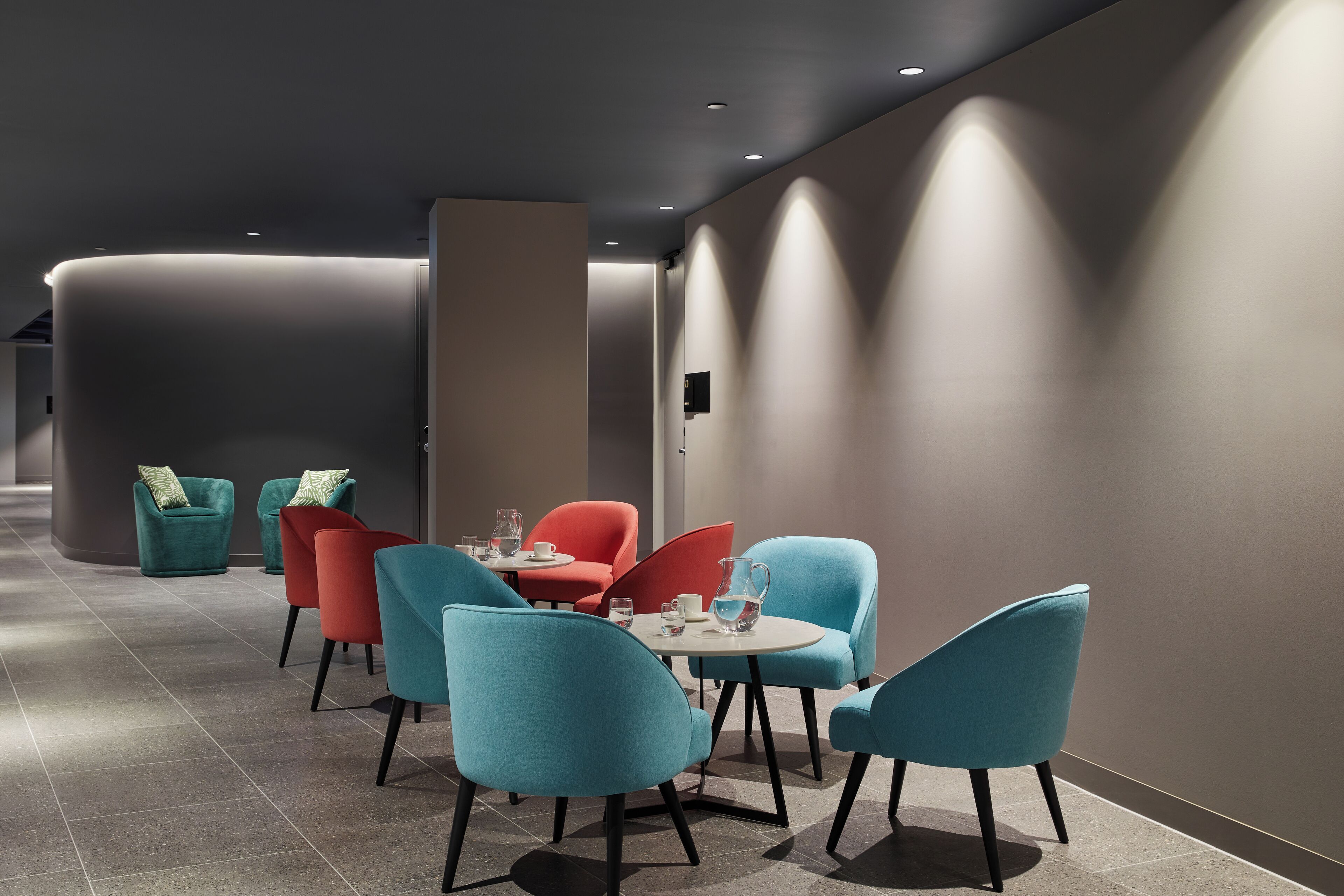 A modern lounge area with teal and red chairs, a round table, and a water pitcher.