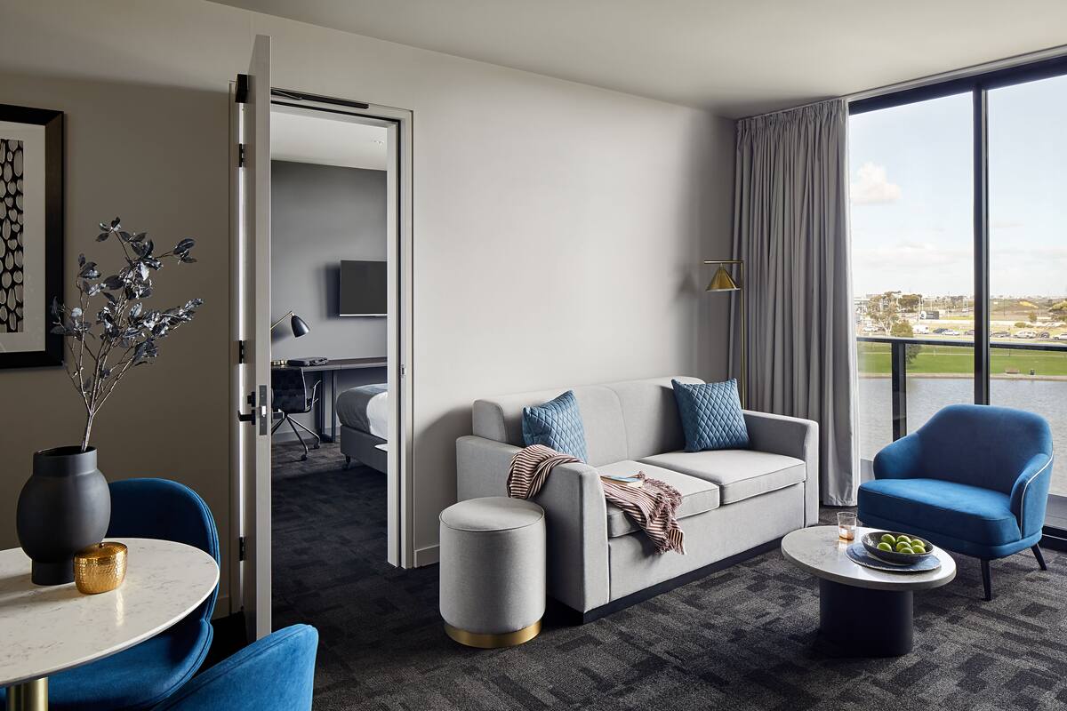 A modern hotel room with a grey sofa, blue armchair, and a small round table with a bowl of fruit.