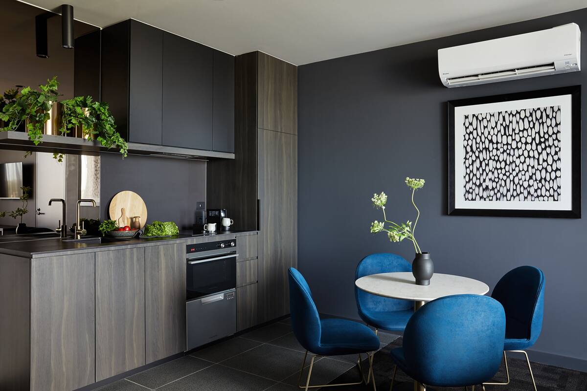A modern kitchen with dark cabinetry, a built-in oven, and a dining area with blue chairs and a round table.